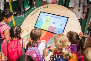 Mehrere Kinder stehen vor dem interaktiven Ausstellungsexponat "Klima-Tourer" und schauen auf ein Display, das die Emissionen unterschiedlicher Transportmittel auf Kurz- und Langstrecken verdeutlicht.