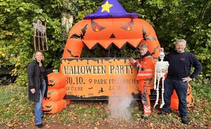 Mitarbeiter der Stadt und der stellvertretende Ortsbrandmeister stehen vor dem Werbebanner für die Halloween-Party