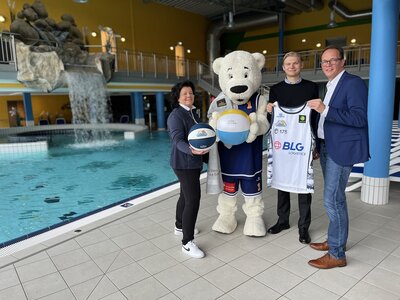 Gabi Kasten, Max Jorzyk und Manuel Kuhn stehen mit dem Maskottchen Dunky neben dem Schwimmbecken in der Moor-Therme